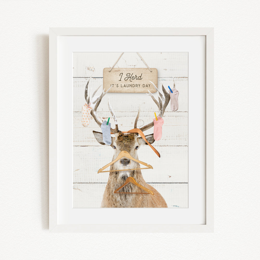 A framed image depicting a deer with antlers holding various laundry items, including socks and clothes, with a sign above that says "I herd its laundry day".