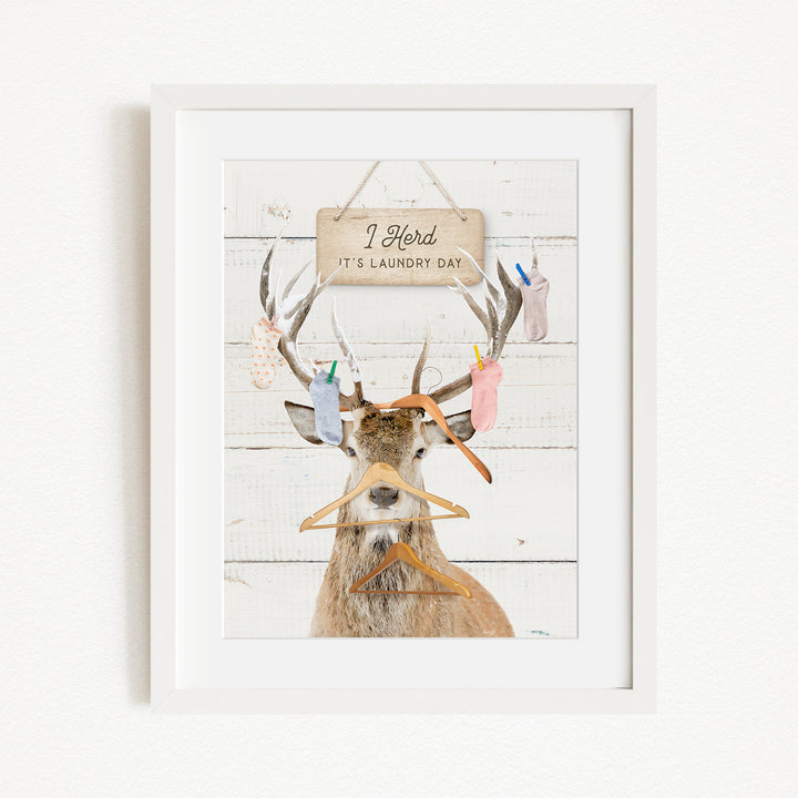 A framed image depicting a deer with antlers holding various laundry items, including socks and clothes, with a sign above that says "I herd its laundry day".
