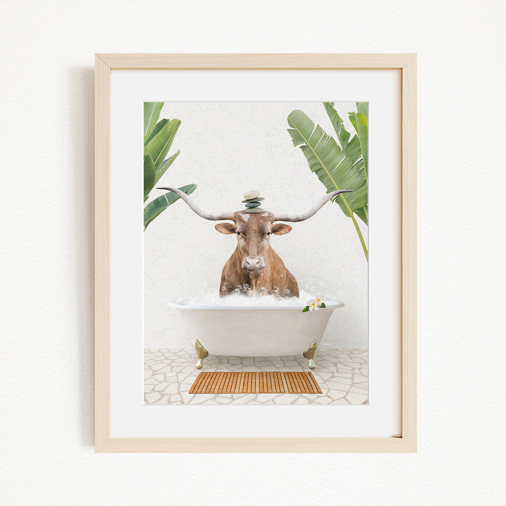 A framed image depicting a brown cow with large horns sitting in a white bathtub filled with water, surrounded by green plants.