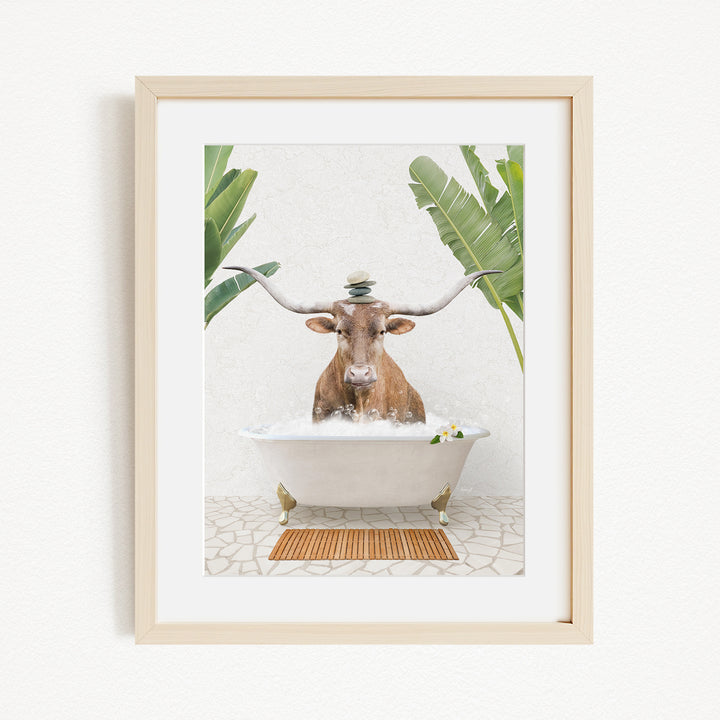 A framed image depicting a brown cow with large horns sitting in a white bathtub filled with water, surrounded by green plants.