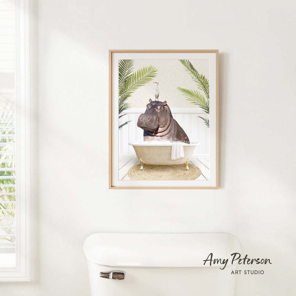 A framed image of a hippopotamus sitting in a bathtub, surrounded by palm leaves, is hanging on a white wall.