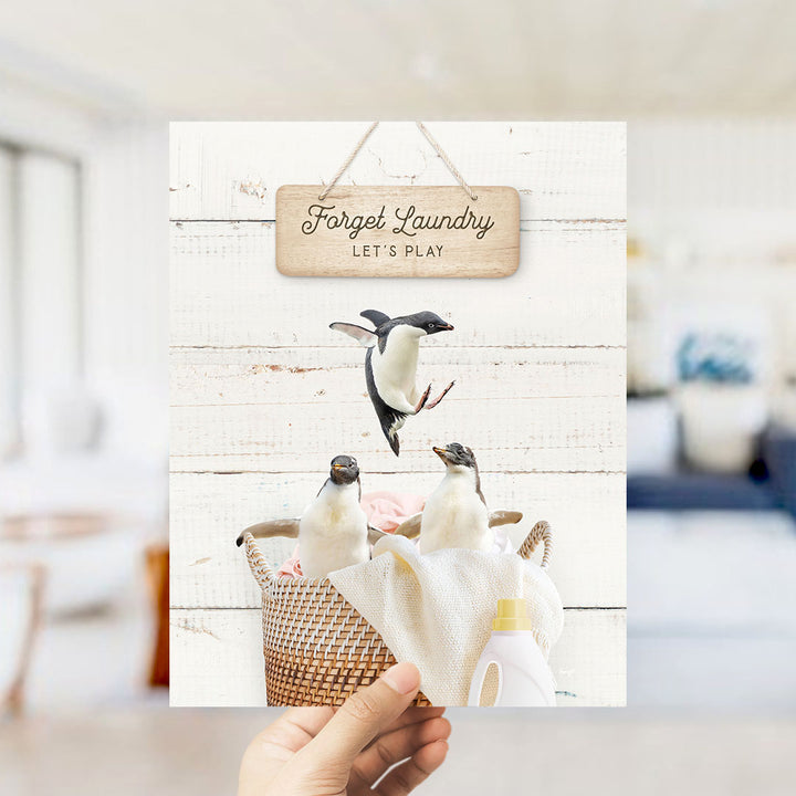 A hand holds up a framed photograph of three penguins in a basket, with a sign that says "Forget Laundry Let's Play" above the image.