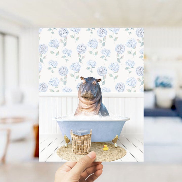 A hand holds up a framed image of a hippopotamus sitting in a bathtub filled with water, surrounded by a blue floral pattern.