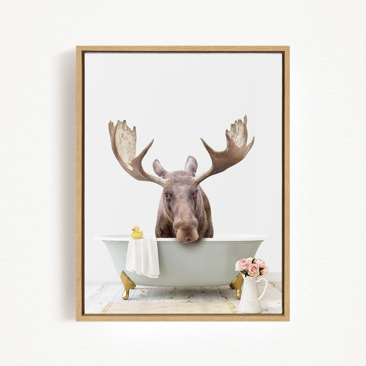 A moose with large antlers is sitting in a bathtub, surrounded by a yellow rubber duck, a white towel, and a vase of pink flowers.