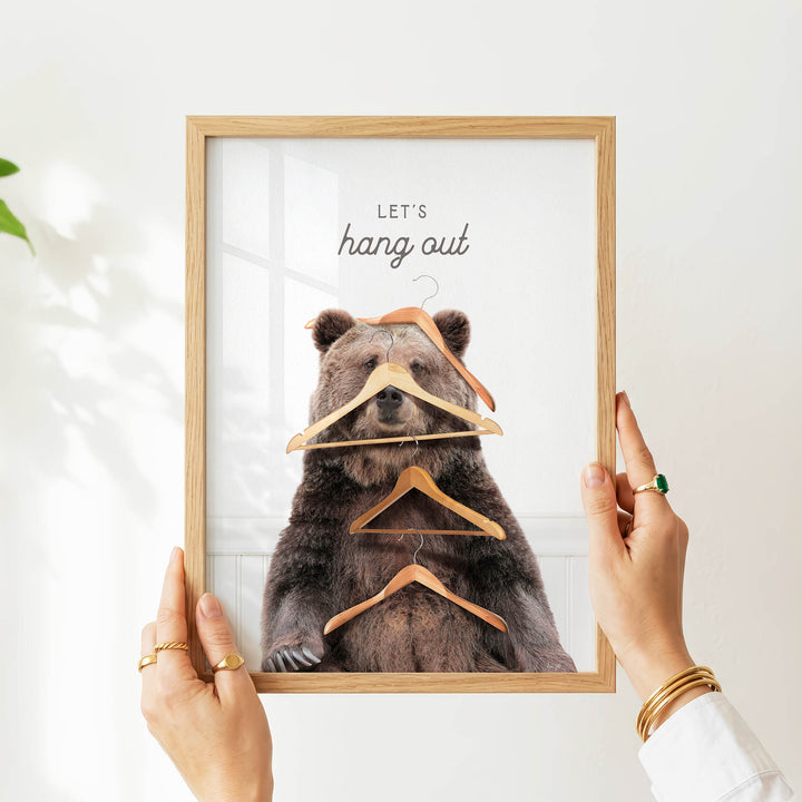 A person's hands are holding a framed picture of a bear with wooden hangers hanging from its head and neck.