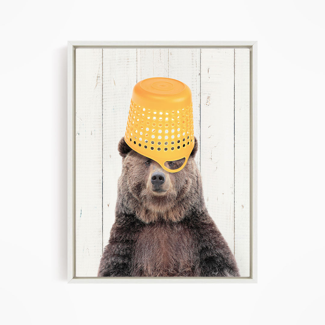A brown bear wearing a yellow plastic bucket on its head, with a white wooden background.