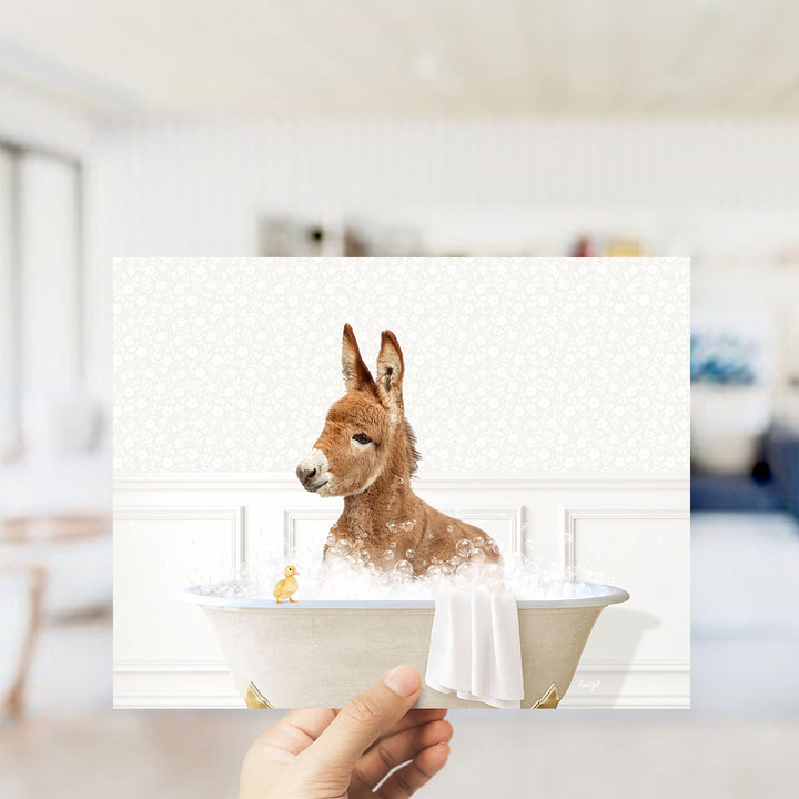 A hand holding a framed image of a brown donkey sitting in a bathtub, surrounded by bubbles, with a small yellow duck nearby.