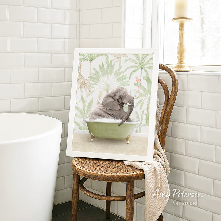 A framed image of a gray elephant sitting in a green bathtub, surrounded by tropical foliage, is displayed on a wooden chair in a bathroom setting.