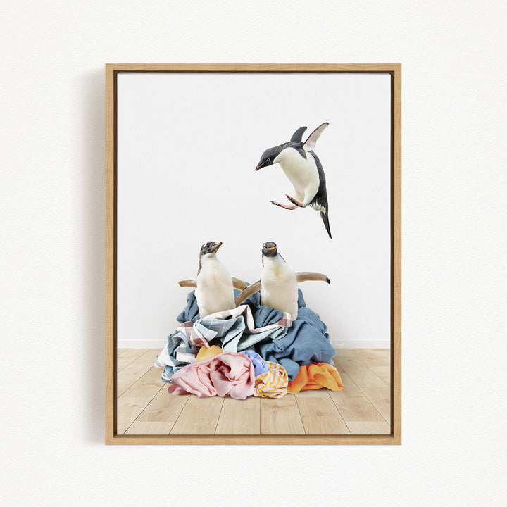 A framed image depicting two penguins standing on a pile of clothes, with one penguin flying in the air above the pile.