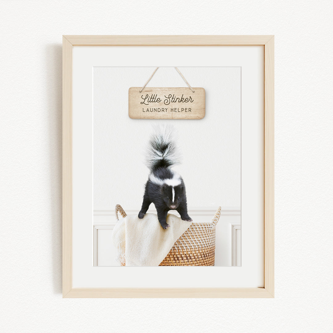 A framed photograph of a black and white skunk standing on a woven basket, with a wooden sign above it that reads "Little Slicker Laundry Helper".