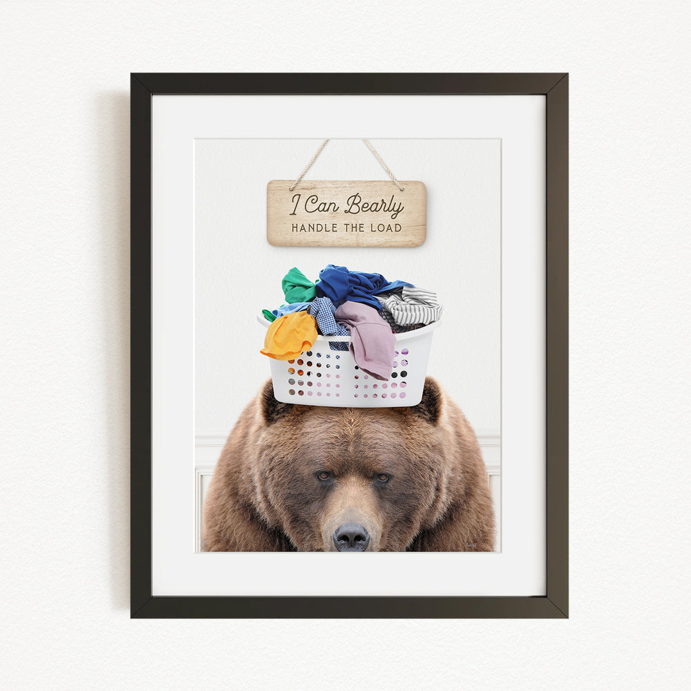 A framed image of a brown bear sitting on top of a laundry basket filled with clothes, with a wooden sign above the bear that says "I Can Bearly Handle the Load".
