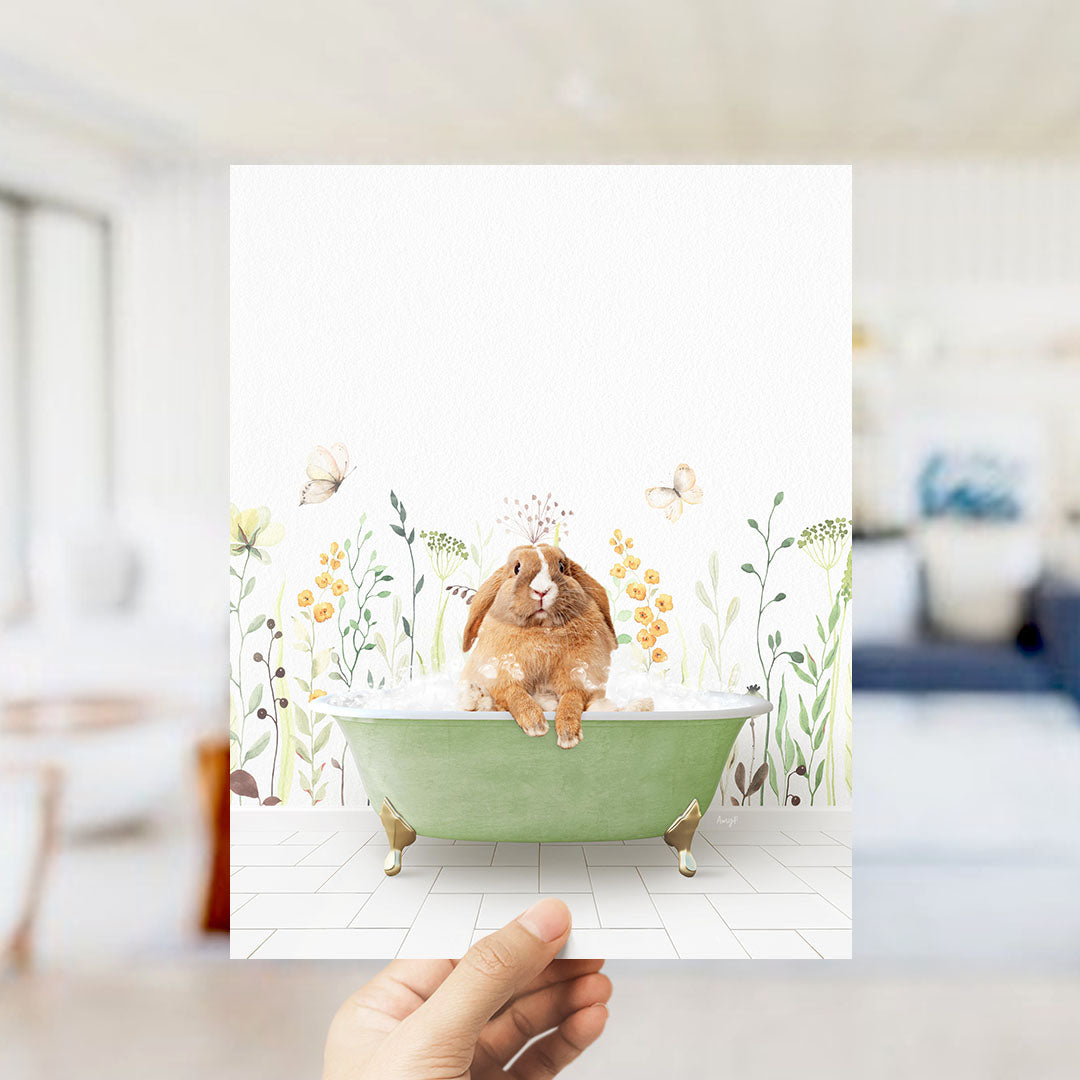 A hand holds up a framed image depicting a brown rabbit sitting in a green bathtub filled with bubbles, surrounded by flowers and butterflies.