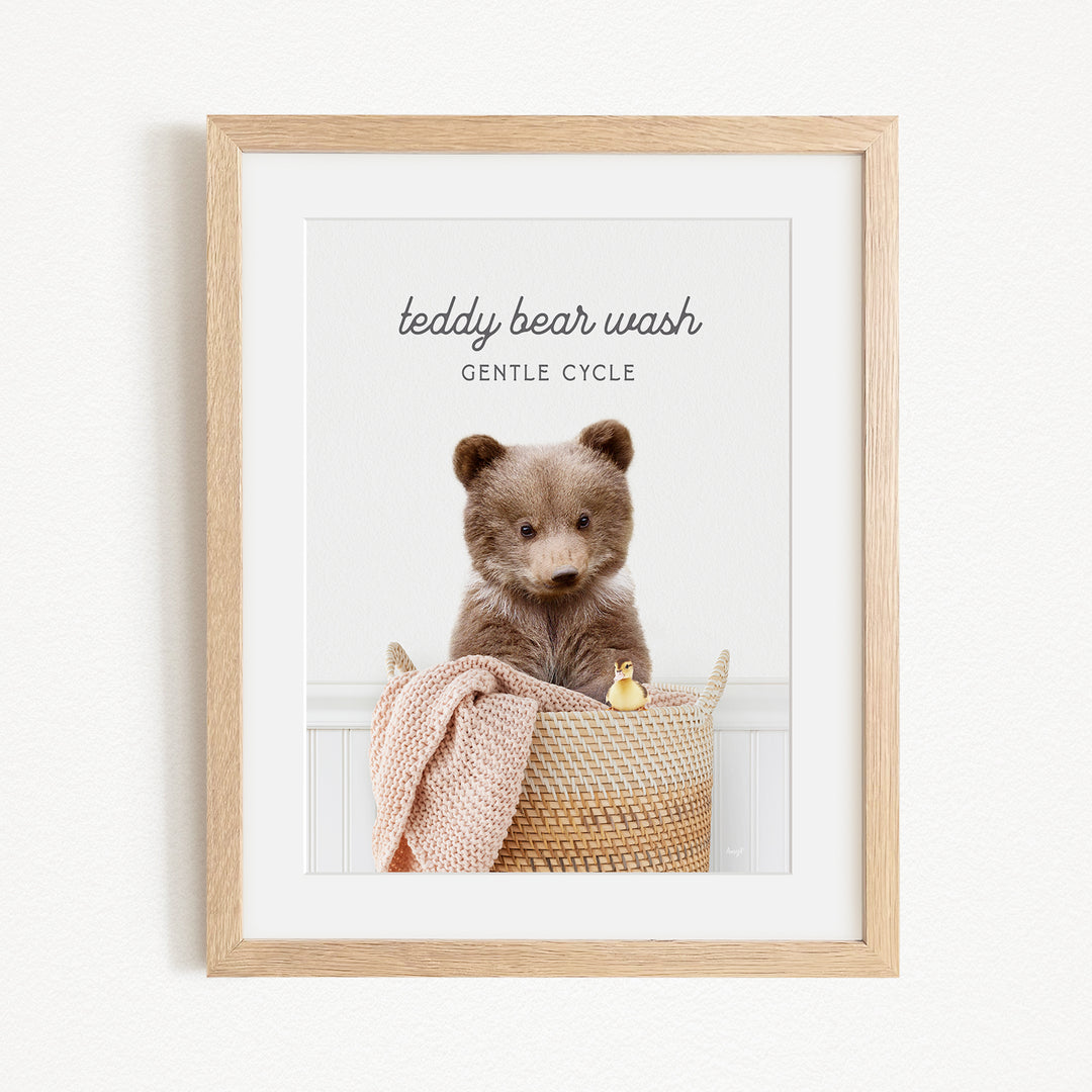 A framed image of a teddy bear sitting in a woven basket, with a blanket and a small toy duck nearby.