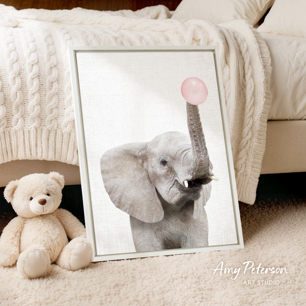 A framed photograph of an elephant holding a pink bubble is displayed on a beige carpet, accompanied by a teddy bear.