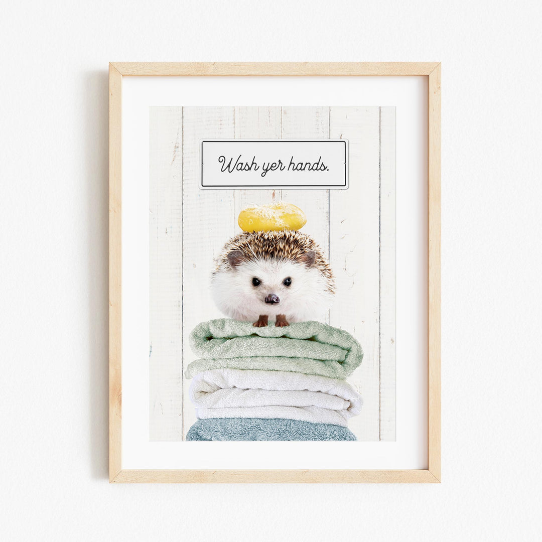 A framed image of a hedgehog sitting atop a stack of folded towels, with the text "Wash your hands" written above it.