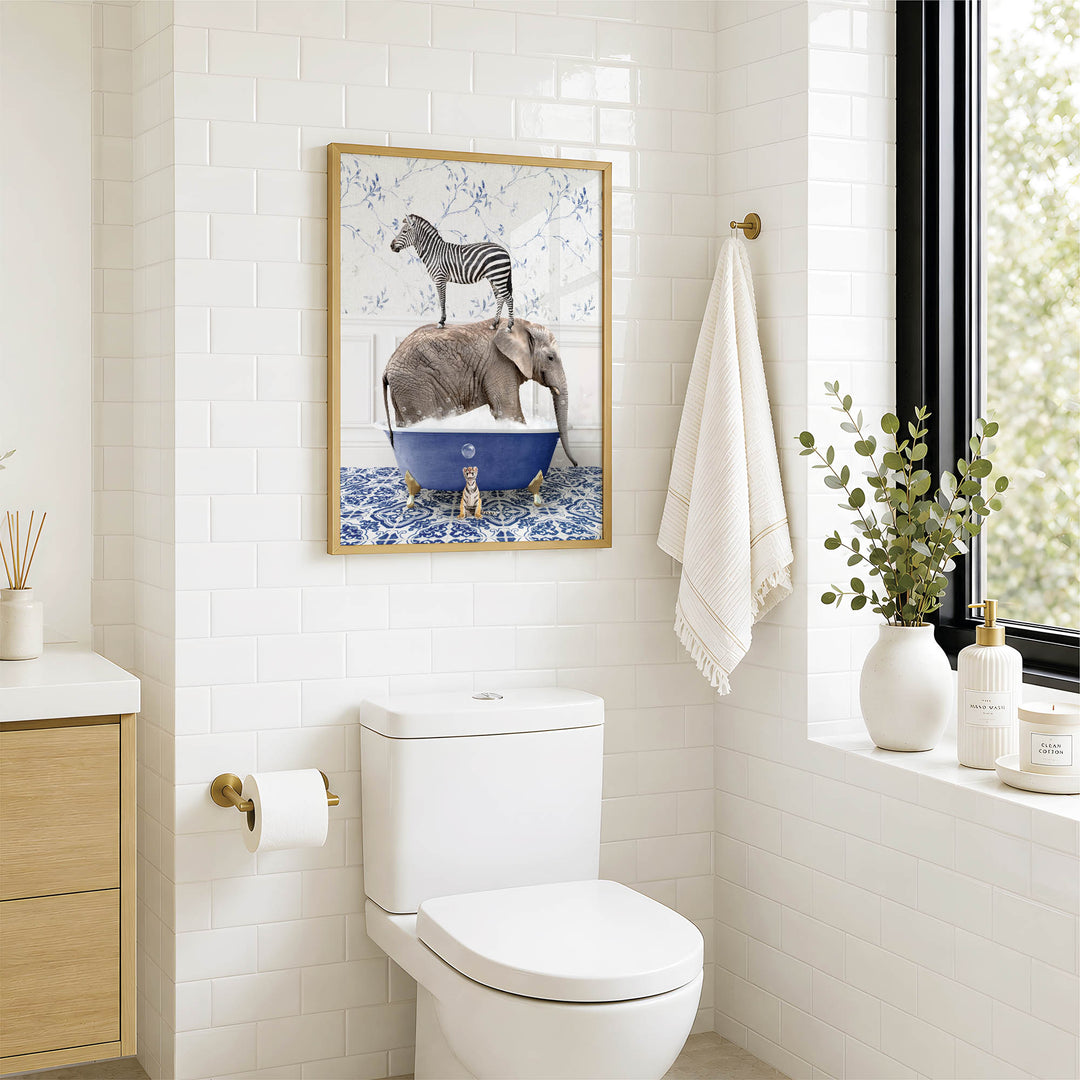 A bathroom with a white toilet, a framed picture of a zebra and an elephant sitting in a blue bathtub, and a window with greenery outside.