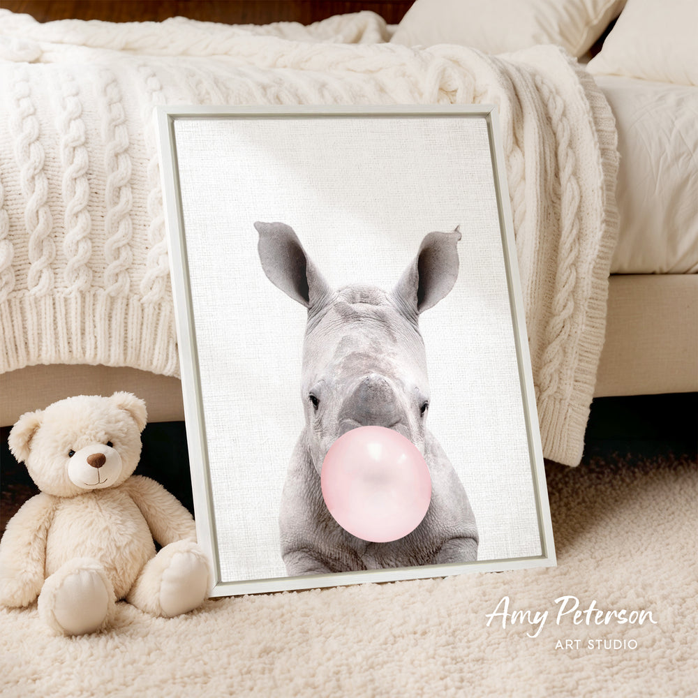 A framed image of a rhinoceros blowing a bubble with a pink bubblegum bubble is displayed on a bed, accompanied by a teddy bear.