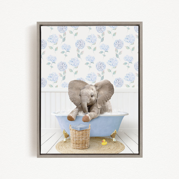 A gray elephant sitting in a blue bathtub, surrounded by a wicker basket and a yellow rubber duck, with a floral patterned wall behind it.