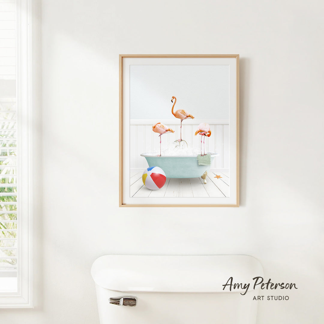 A framed print of a vibrant, colorful scene depicting a bathtub filled with three flamingos, a beach ball, and a towel, hanging on a white wall.
