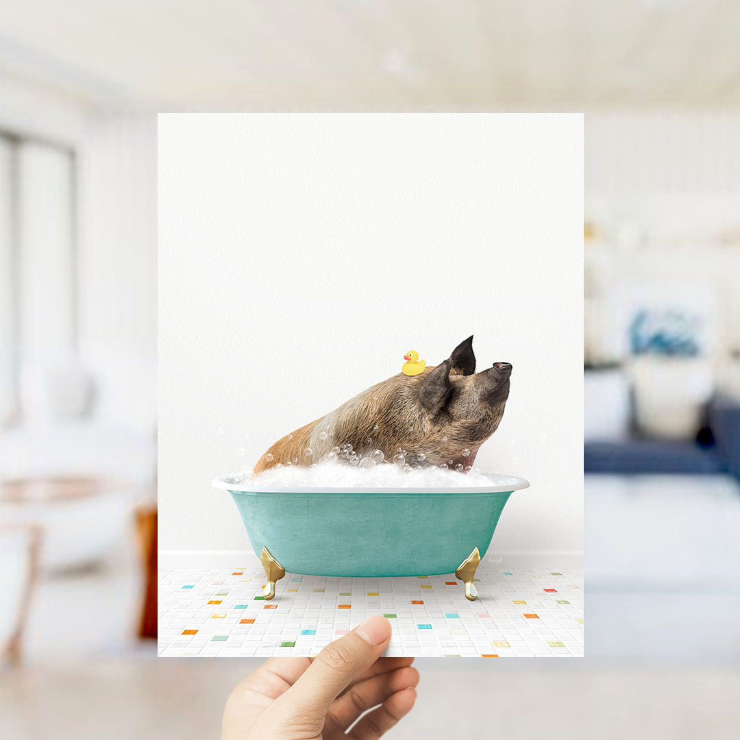 A hand holds up a framed image of a pig sitting in a bathtub filled with bubbles, with a yellow rubber duck floating on its head.