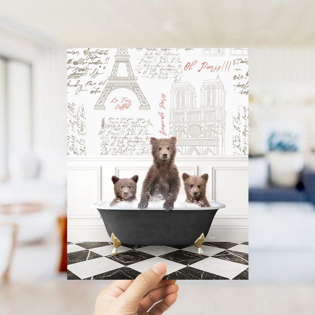 A hand is holding up a framed image depicting three bears sitting in a bathtub, with the Eiffel Tower and Notre-Dame Cathedral visible in the background.