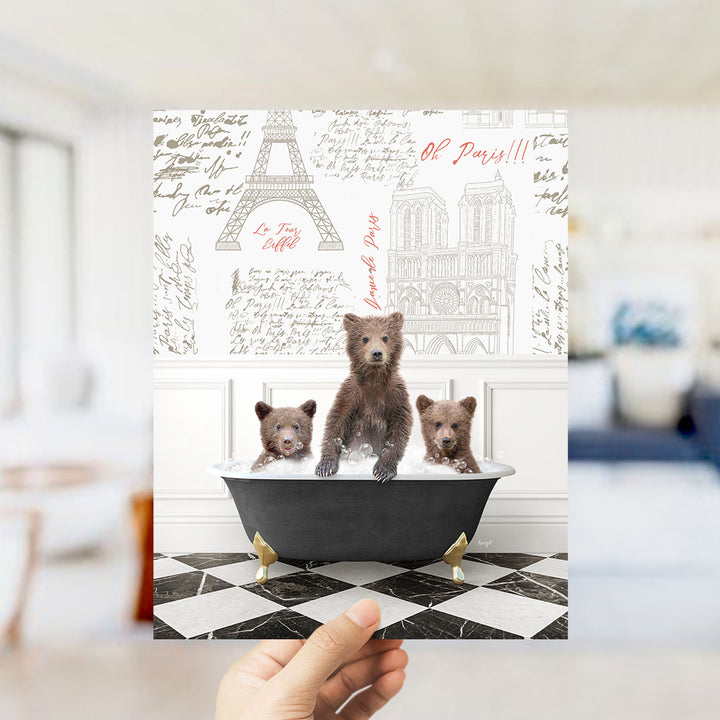 A hand is holding up a framed image depicting three bears sitting in a bathtub, with the Eiffel Tower and Notre-Dame Cathedral visible in the background.