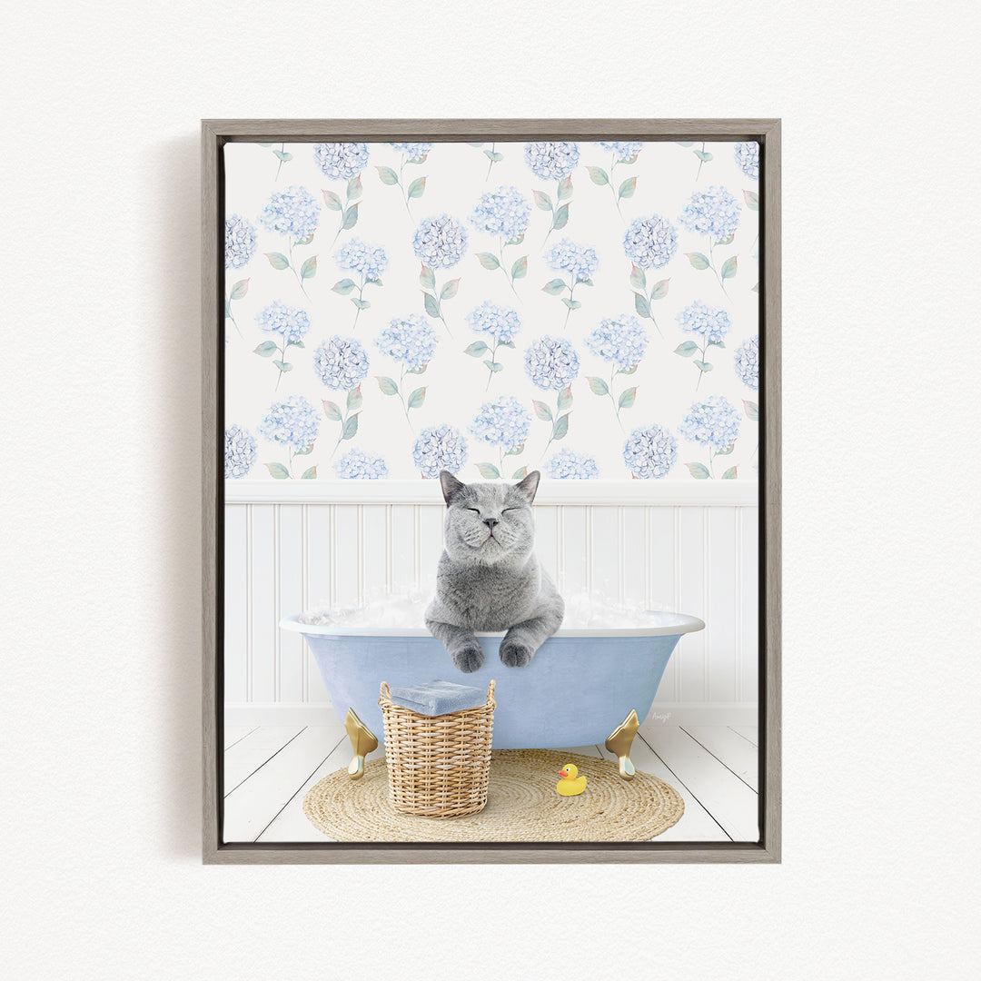 A gray cat sitting in a blue bathtub, surrounded by a wicker basket, a rubber duck, and a floral patterned wall behind it.