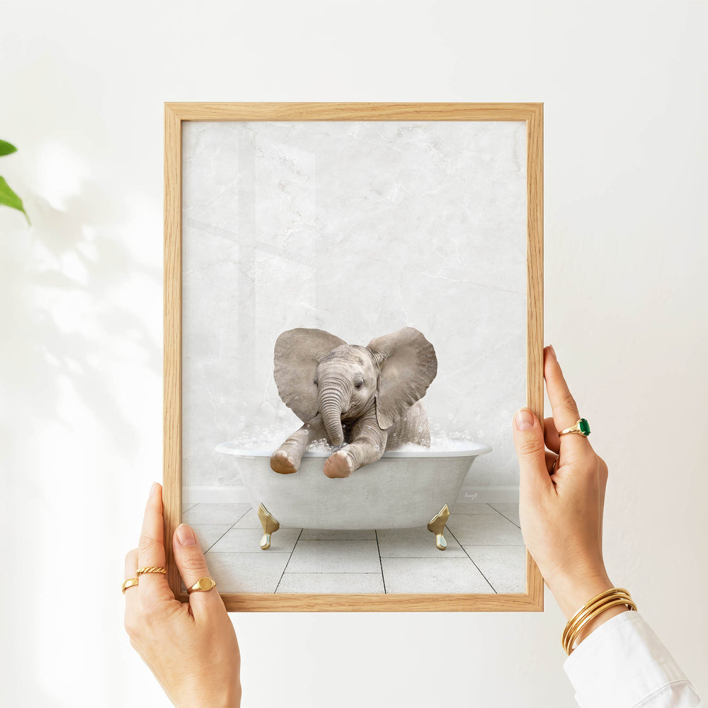 A person's hands are holding a framed picture of a baby elephant sitting in a bathtub.