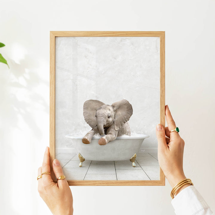 A person's hands are holding a framed picture of a baby elephant sitting in a bathtub.