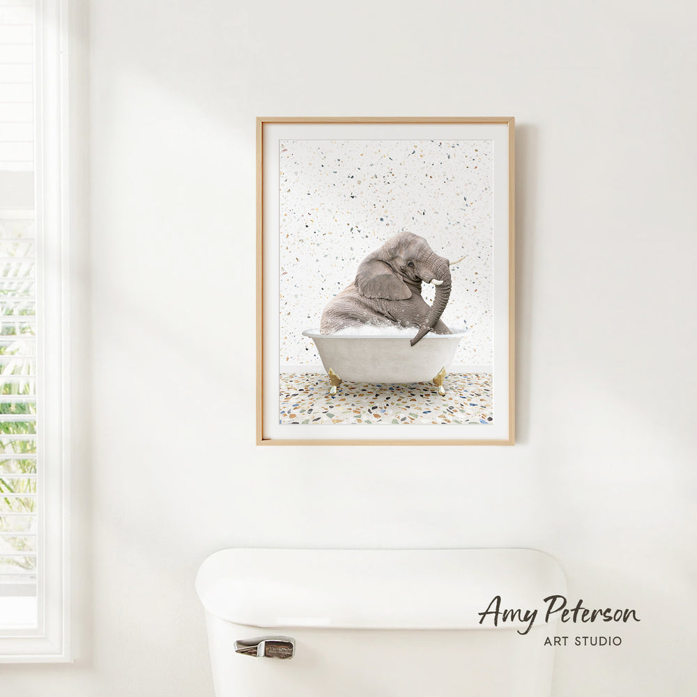 A framed image of a gray elephant sitting in a white bathtub, surrounded by multicolored confetti, is hanging on a white wall.
