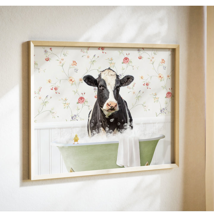 A framed image depicting a black and white cow sitting in a green bathtub filled with bubbles, with a yellow bird perched on the edge of the tub.