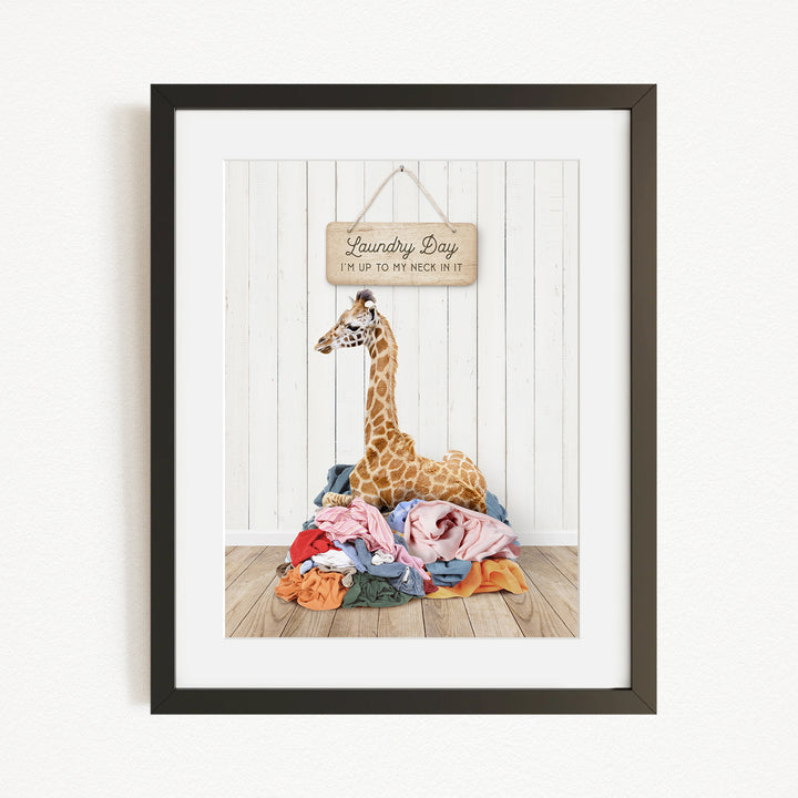 A giraffe is sitting on a pile of clothes in front of a sign that says "Laundry Day"