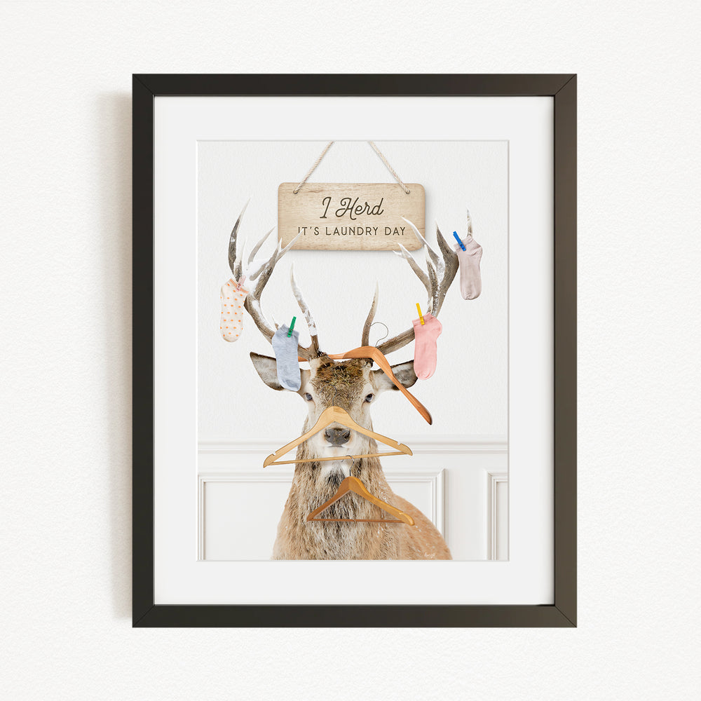 A framed image depicting a deer with antlers holding various laundry items, including socks and clothes hangers, with a sign above that says "I Harded It's Laundry Day".