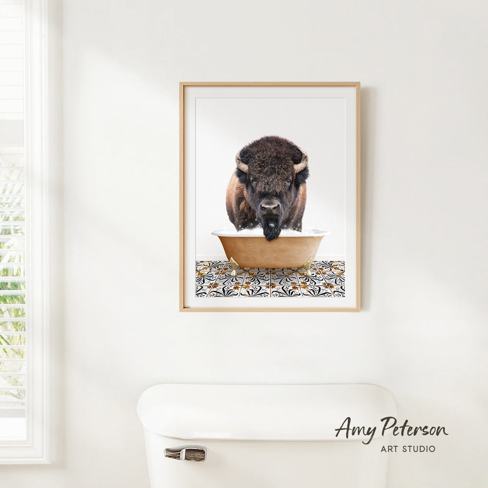 A framed image of a buffalo standing in a bowl, with a patterned mat below it, is hanging on a white wall.