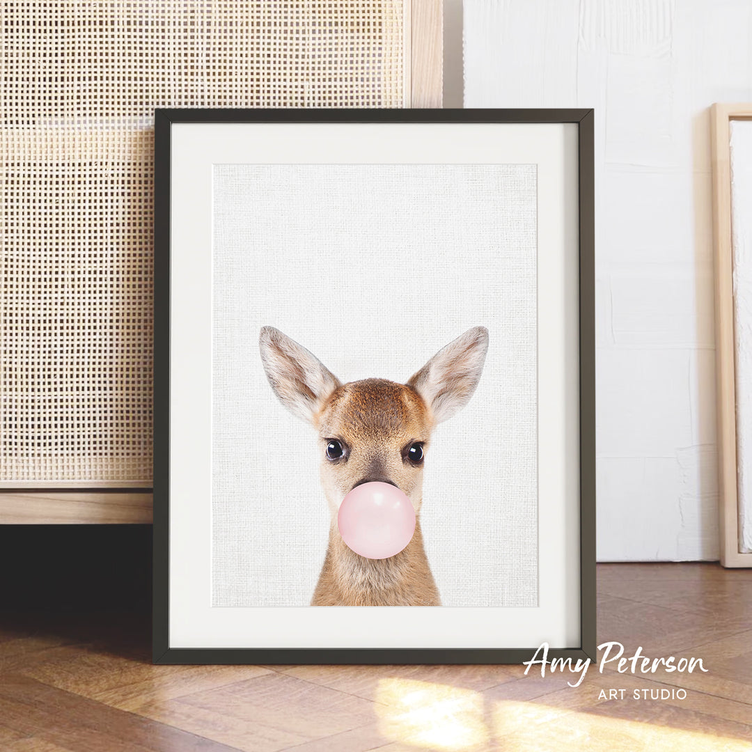 A framed print of a deer blowing a bubble gum bubble, with a wooden floor and a woven basket in the background.