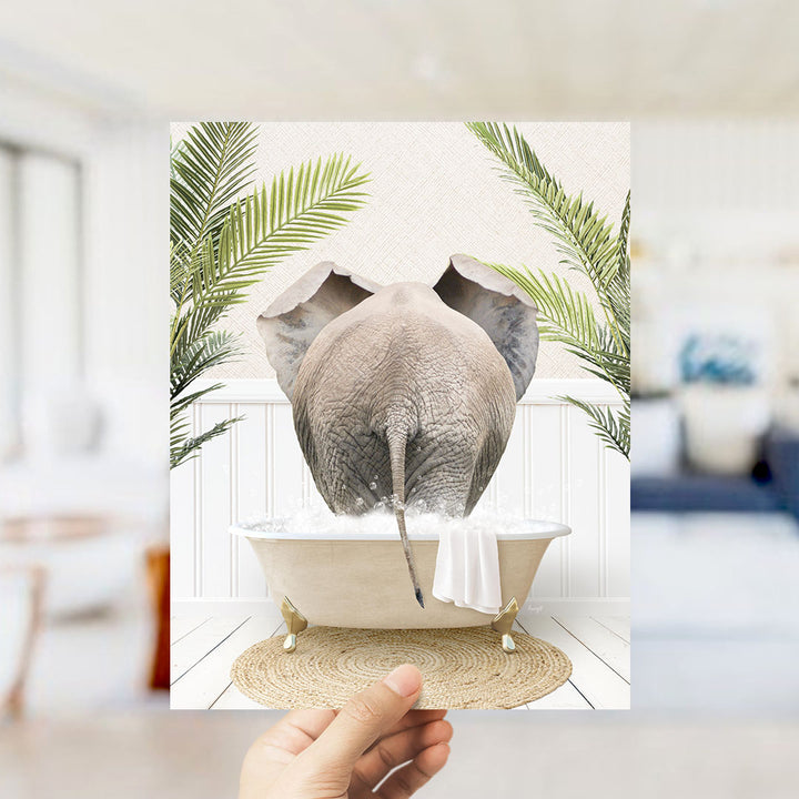 A hand holds up a framed image depicting an elephant sitting in a bathtub filled with water, surrounded by palm leaves.