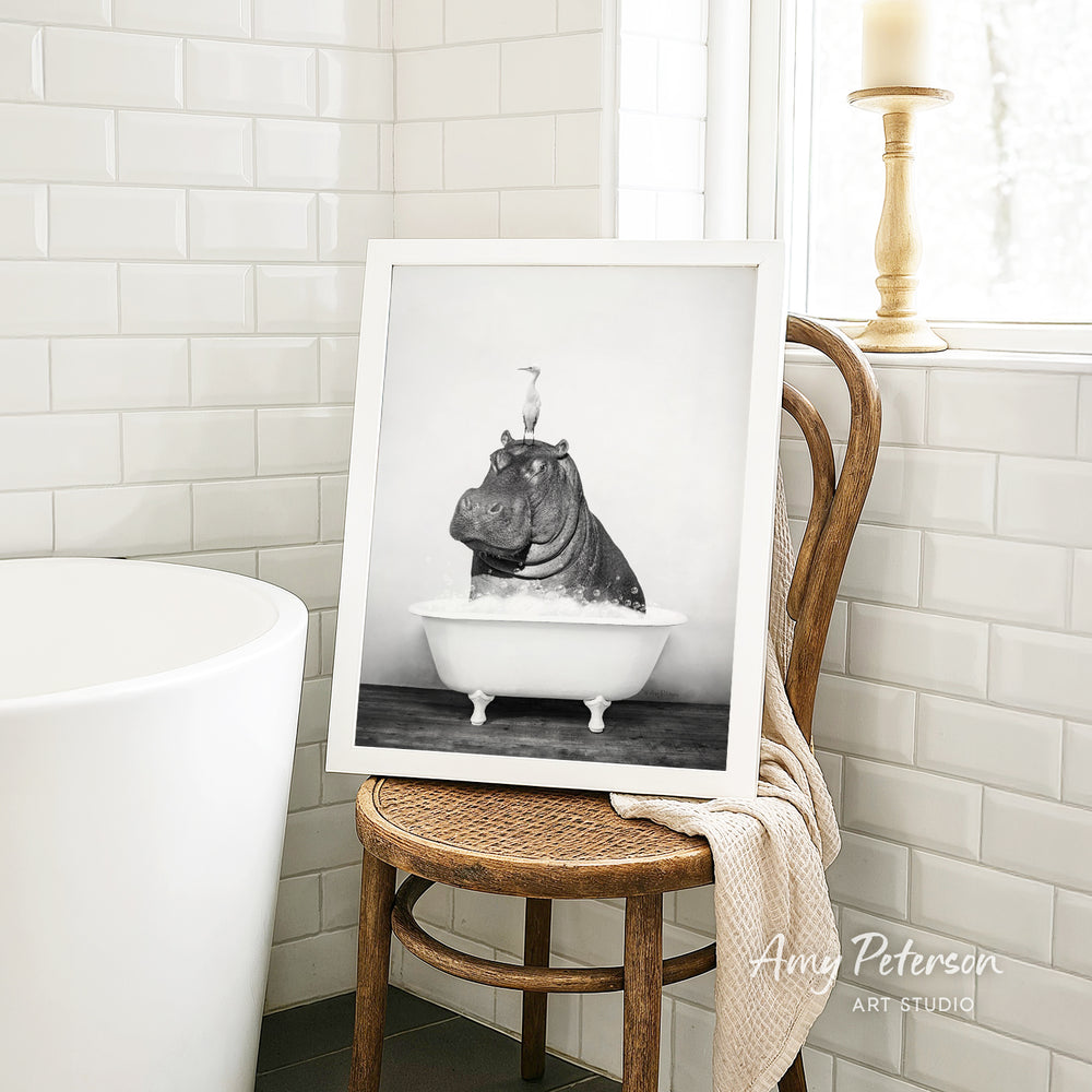A framed black and white photograph of a hippopotamus sitting in a bathtub is displayed on a wooden chair in a bathroom setting.