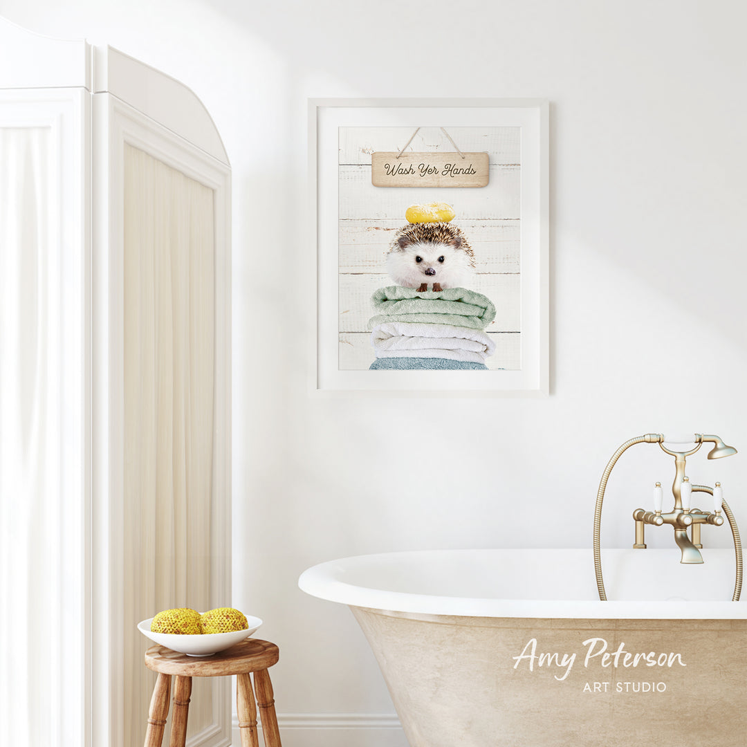 a bathroom with a framed picture of a hedgehog sitting on a stack of towels hanging on the wall. The bathroom has a clawfoot bathtub, a wooden stool, and a bowl of fruit on the floor.