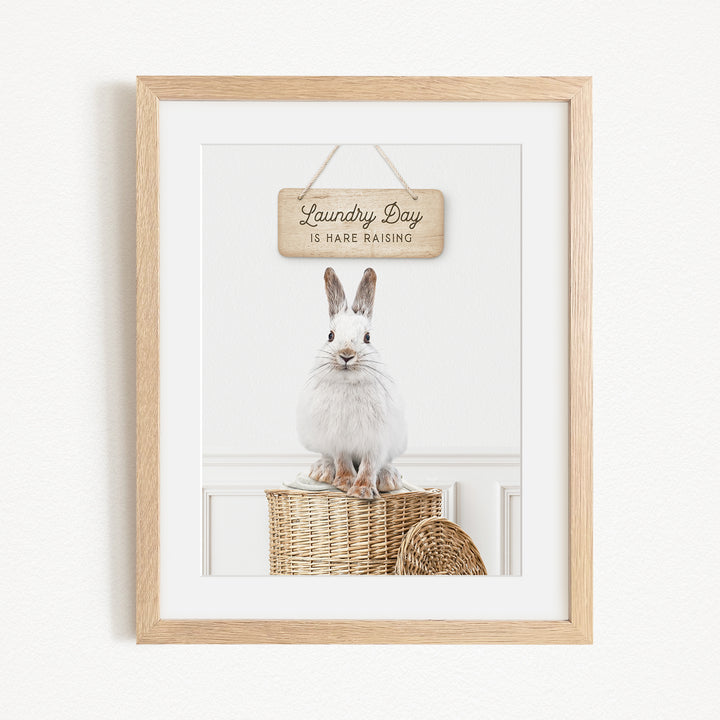 A framed image of a white rabbit sitting on a wicker basket, with a wooden sign above it that says "Laundry Day is here raising".