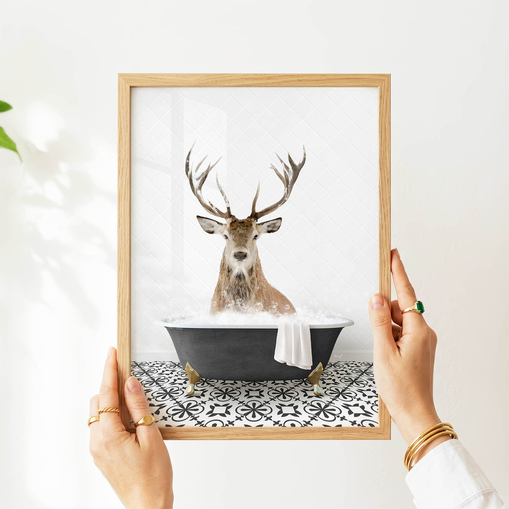 A person's hands are holding a framed picture of a deer sitting in a bathtub filled with water, with a towel hanging from the side of the tub.