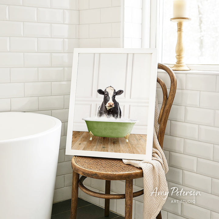 A framed image of a cow sitting in a green bathtub, surrounded by white tiles and a wooden chair.