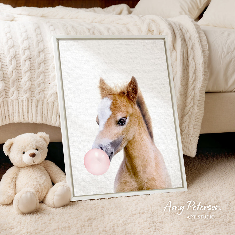A framed image of a horse blowing a bubble gum bubble is displayed on a bed, accompanied by a teddy bear.