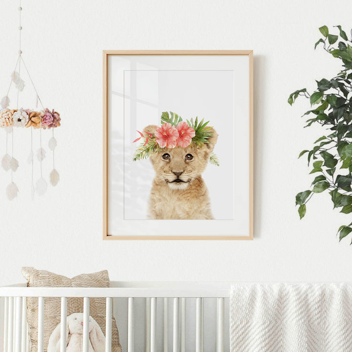 A framed picture of a young lion wearing a floral crown is displayed on a white wall in a nursery setting.