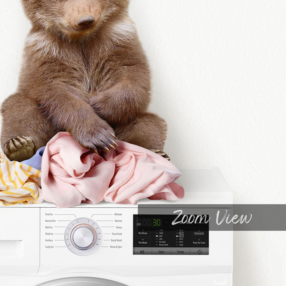 A brown bear sitting on a pile of clothes next to a washing machine.