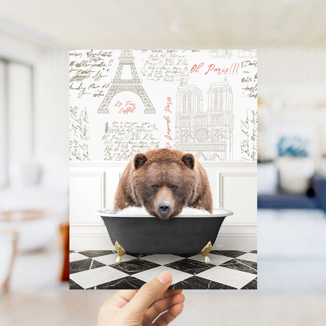A hand holds up a collage featuring a brown bear sitting in a bathtub, with the Eiffel Tower and Notre-Dame Cathedral visible in the background.