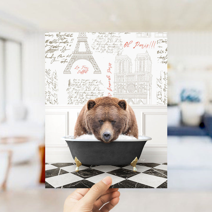 A hand holds up a collage featuring a brown bear sitting in a bathtub, with the Eiffel Tower and Notre-Dame Cathedral visible in the background.