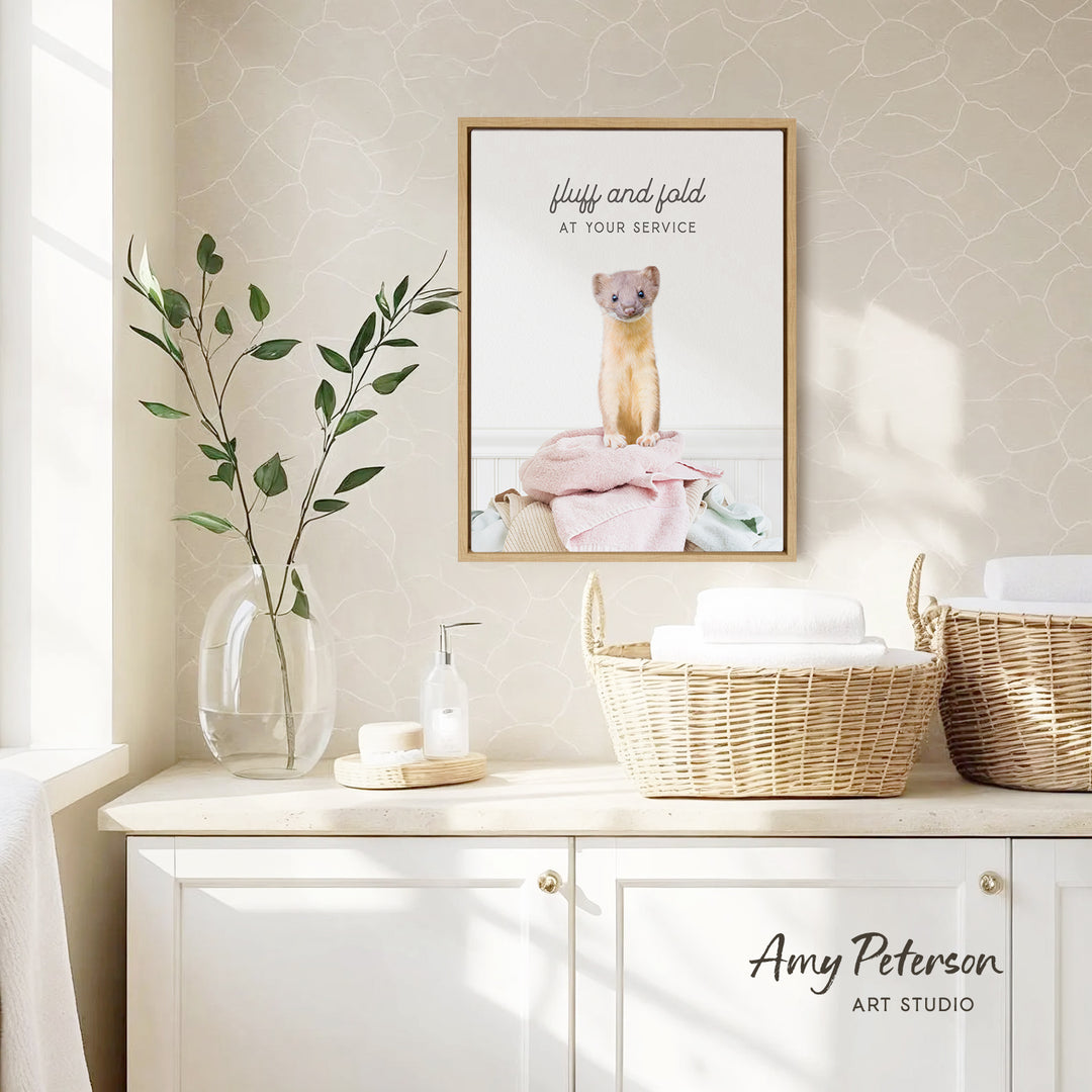 a framed poster of a small animal, likely a rodent, sitting on a pink towel in a bathroom setting. The poster is hung on a wall above a white cabinet, which has two woven baskets on top. The cabinet also has a soap dispenser and a plant on it. The overall scene suggests a cozy and well-decorated bathroom.