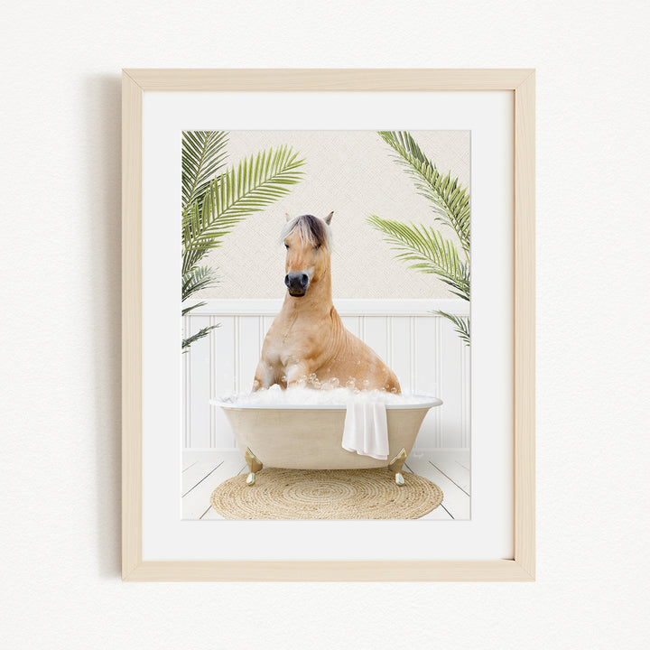 A light brown horse is sitting in a white bathtub filled with water, surrounded by green palm leaves.