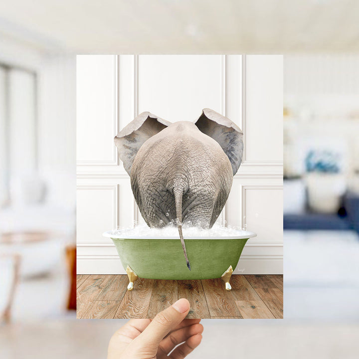 A hand holds up a framed image of an elephant sitting in a green bathtub filled with water, creating a whimsical and playful scene.