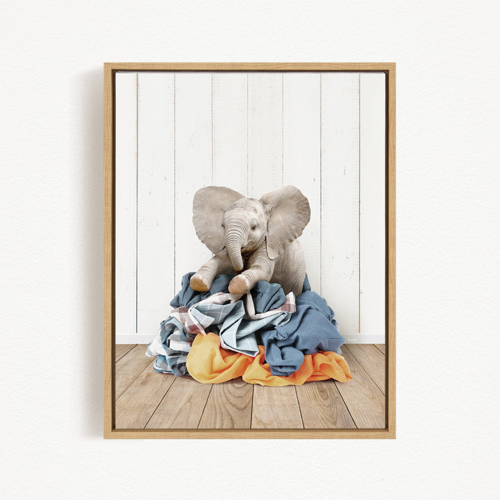 A gray elephant sits atop a pile of clothes on a wooden floor, with a white wall and wooden paneling in the background.